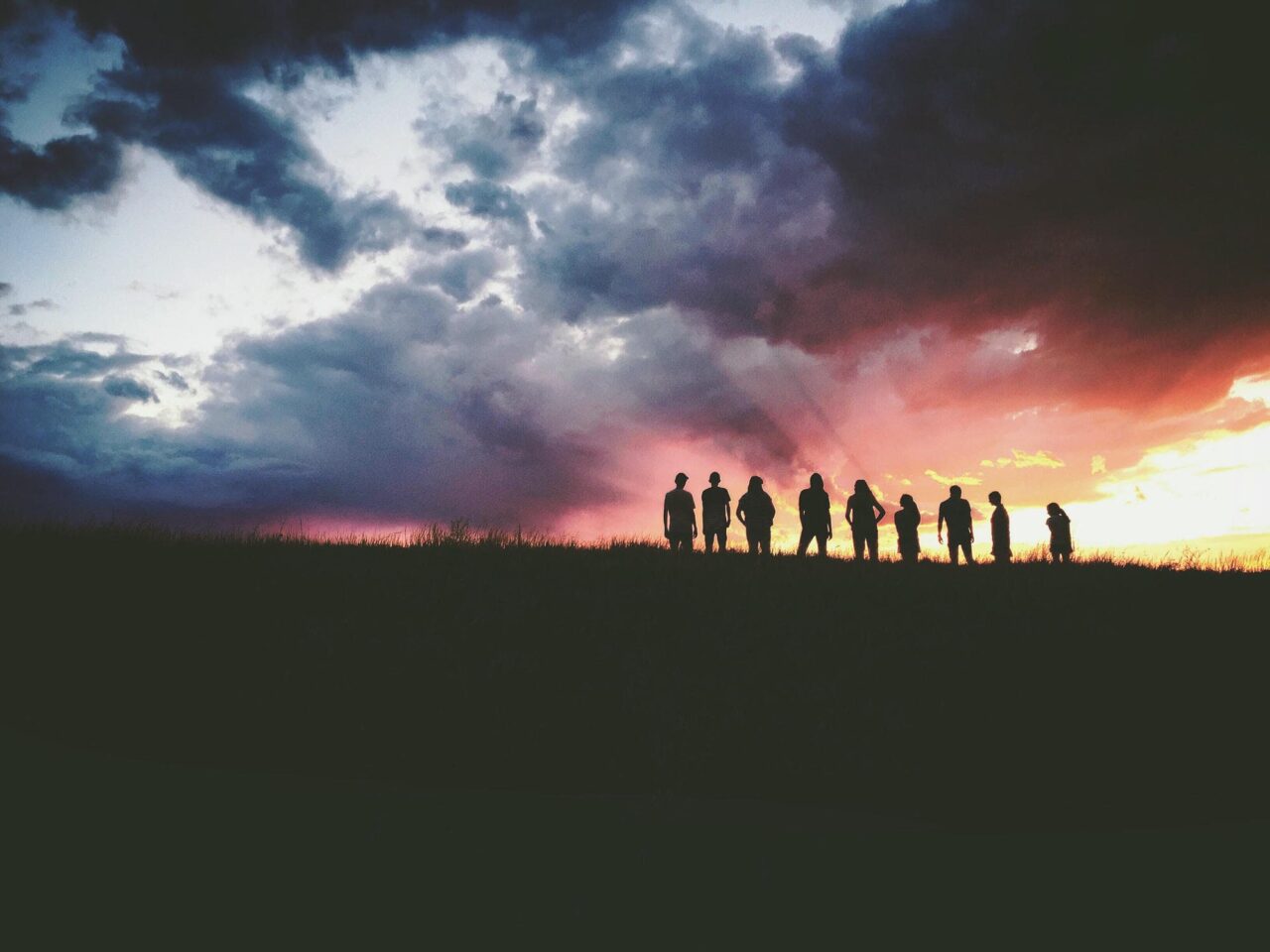 photo of a group of people standing in a field at sunset, the clouds are blue, red and orange and the humans are black silhouettes against the sunset sky